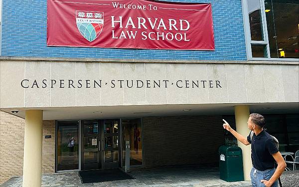 Ein Mann steht unter einem Haus und zeigt auf ein Schild. Auf diesem steht "Welcome to Harvard Law School"