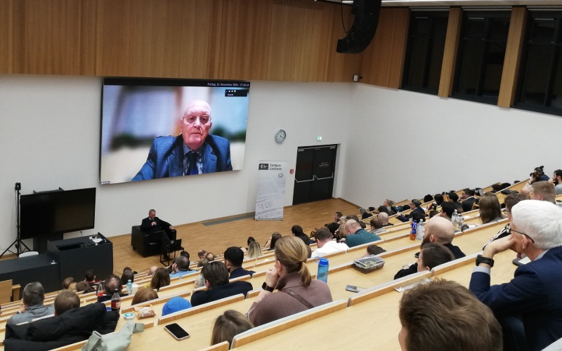 Ein Hörsaal von hinten, vorne auf der Leinwand ist ein Mann zu sehen