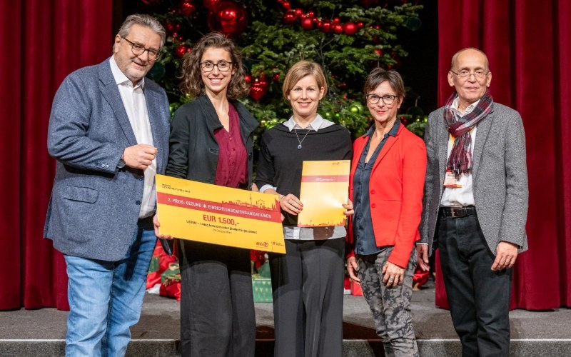 Ein Mann, drei Frauen, ein Mann stehen auf einer Bühne und posieren mit einem Scheck in der Hand