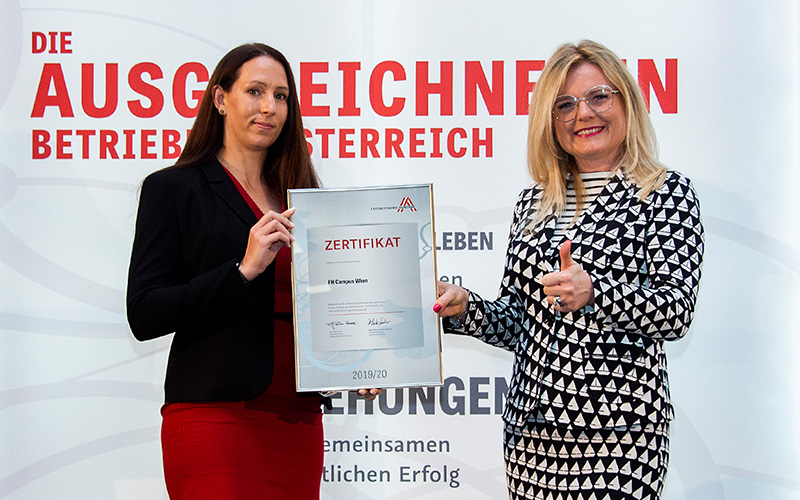 Zwei Frauen stehen vor einem Roll-up auf dem "Die ausgezeichneten Betriebe in Österreich" steht. Links steht Daniela Janko, Qualitätsleitung der FH Campus Wien, rechts steht Monica Rintersbacher, Geschäftsführerin Leitbetriebe Austria